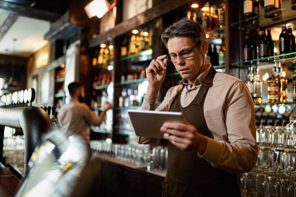 Mid Adult Bartender Using Touchpad While Working In A Pub. - Nexxo Inteligência Empresarial
